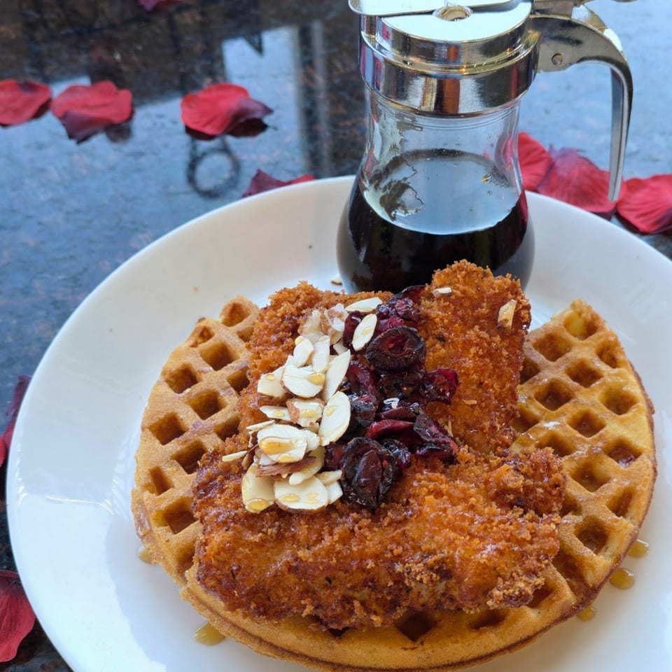 Crispy fried chicken and dried cranberries on a waffle with syrup, on a dark table with rose petals.