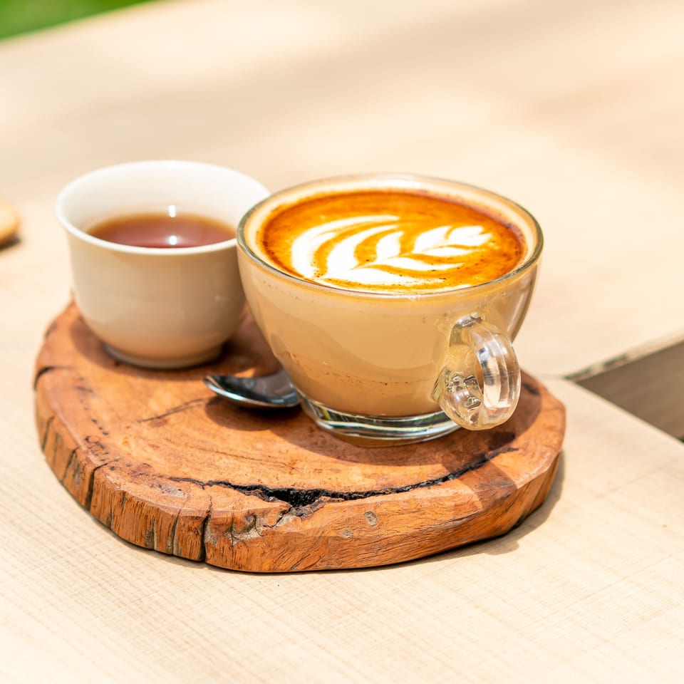 A latte with leaf art and a small cup of tea on a rustic wooden coaster.