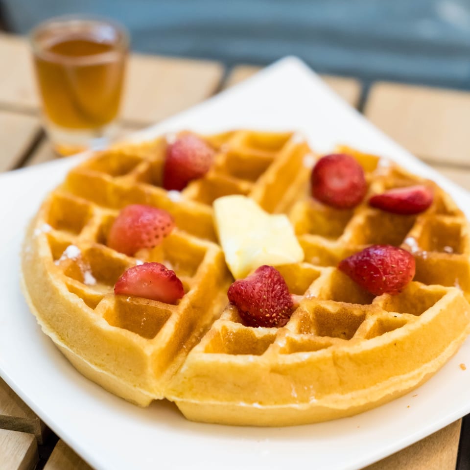 Golden waffle with strawberries, butter, and powdered sugar, with a drink in the background.