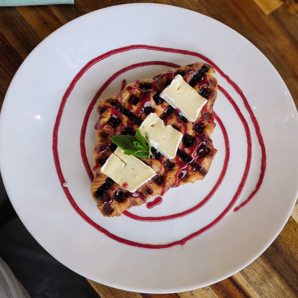 Croffle with berry sauce, brie cheese, and mint on a white plate with red drizzle.